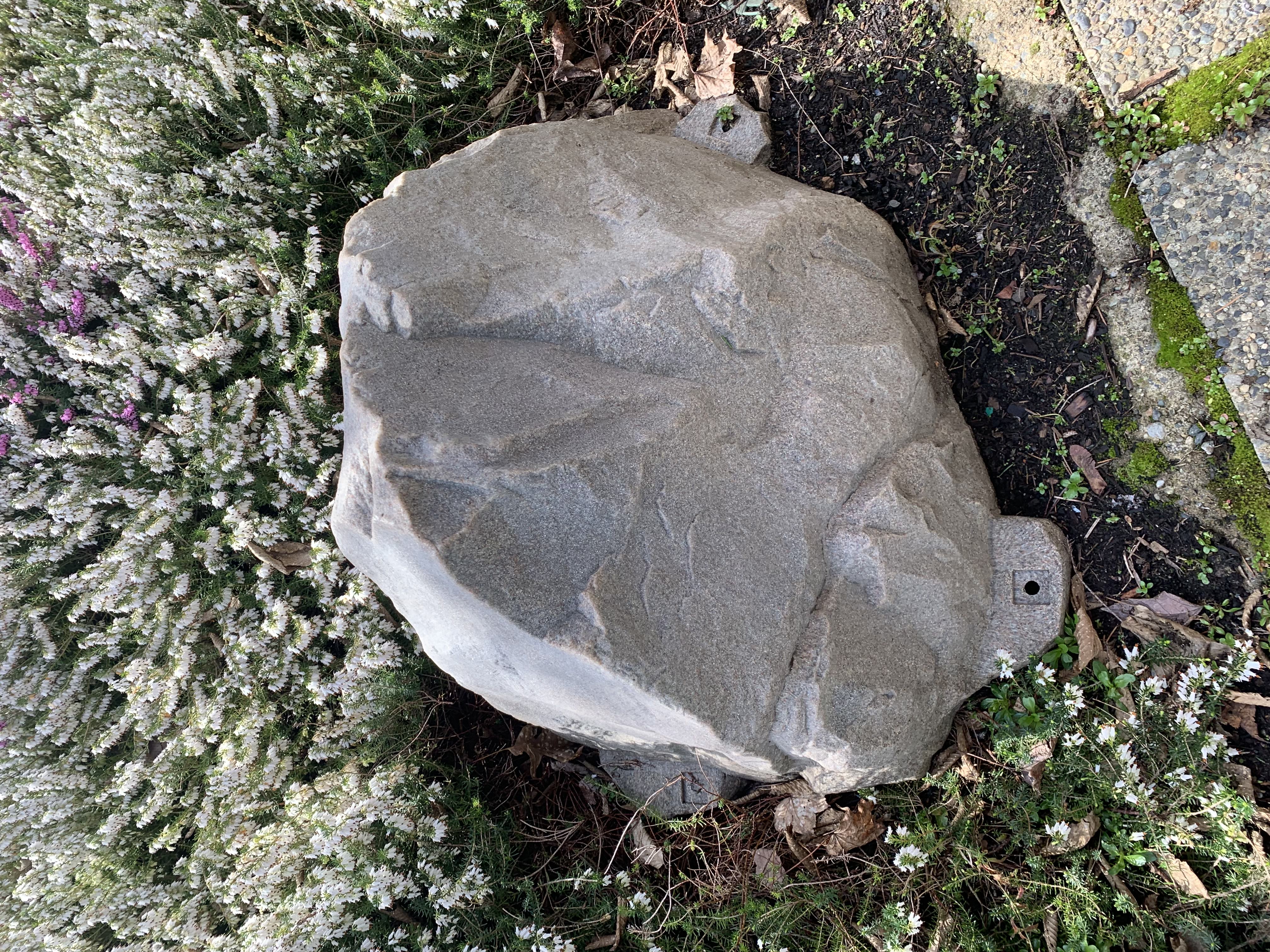 close up of a fake rock with
             exposed screw hole for mounting, sidewalk stones in foreground, hedge surrounding