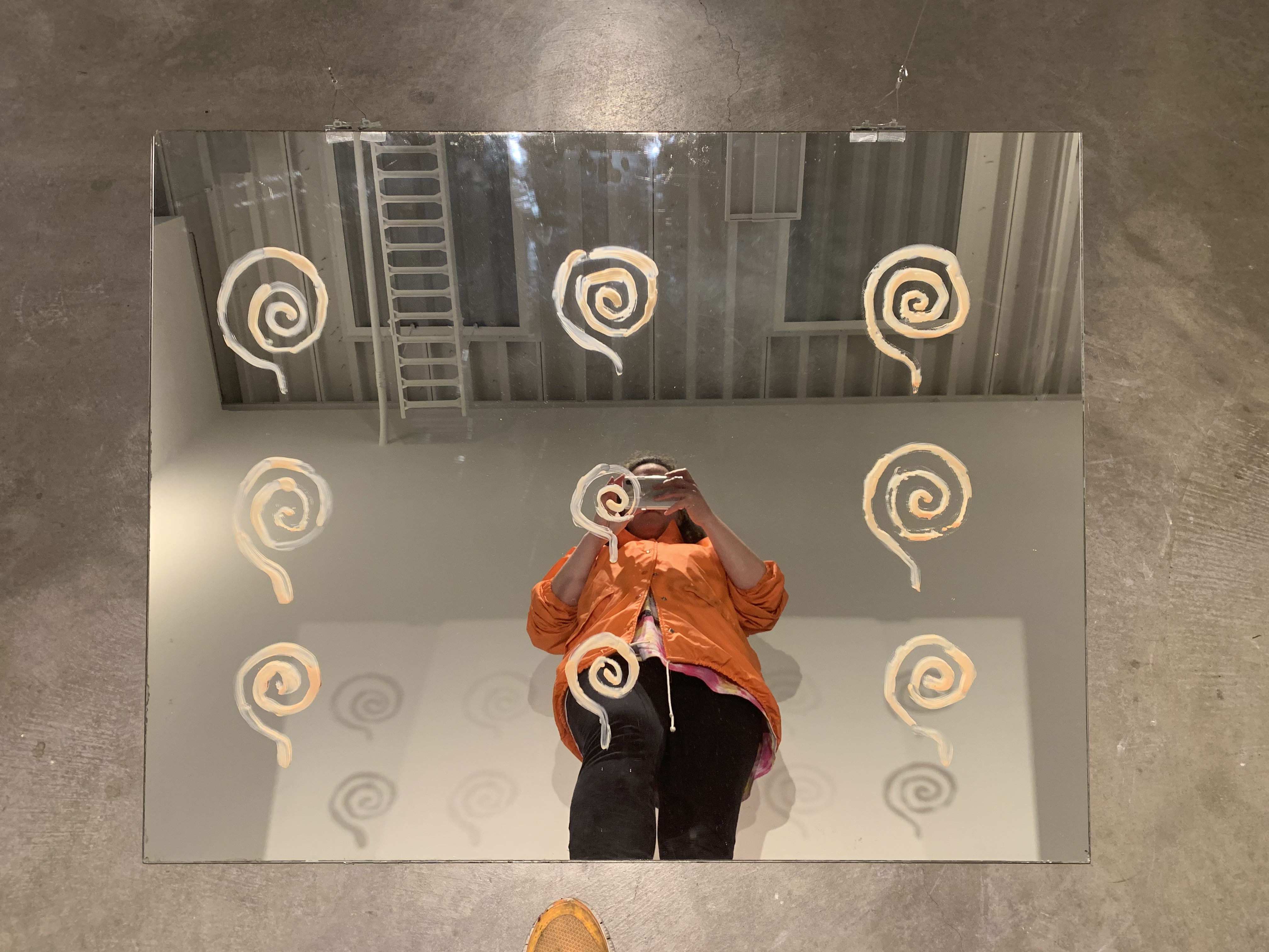 a human looking down at a mirror. the majority of their figure is
             reflected. the face is covered by the phone taking the picture, held with both hands. the large frameless
              mirror sits on the floor at and angle and has nine spirals painted on it. behind the figure the mirror
              reveals an industrial ceiling and light reflecting the spirals on the wall behind the figure. the figure
               wears an orange jacket and bottom frame an orange shoe peeks in to frame.